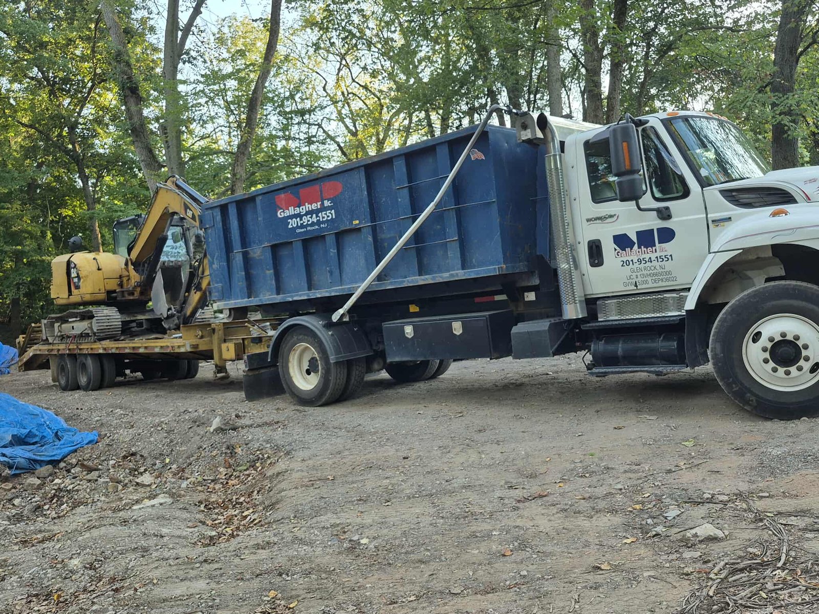 MP Gallagher LLC construction dump truck with blue container and yellow excavator on trailer at a job site, used for excavation, hauling, and debris removal services in New Jersey.